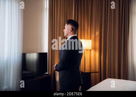 Ein junger Mann in einem Anzug, der in einem Hotelzimmer steht, in das Fenster schaut, meditiert, sich auf einen großen Tag vorbereitet, Rückansicht. Stockfoto