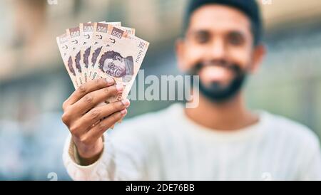 Junger afroamerikanischer Mann lächelt glücklich und hält mexikanische 500 Pesos Banknoten in der Stadt. Stockfoto