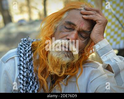 Der alte indische Hindu-Mann mit Henna-gefärbten langen Haaren und langem Bart schläft im Freien bei hellem Tageslicht und legt seine Stirn auf seine linke Hand. Stockfoto