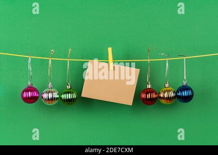 Bunte weihnachtskugeln und unbeschriftete braune Grußkarte auf einem an einer Schnur vor grünem Hintergrund hängenden. Winterferien Weihnachten und Neujahr. Stockfoto