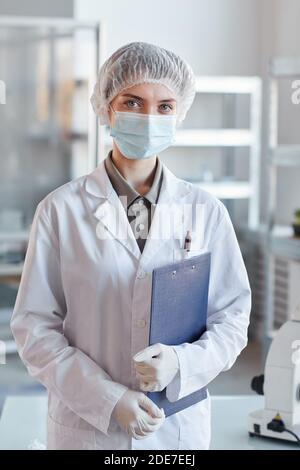 Vertikale Taille nach oben Porträt der jungen Wissenschaftlerin trägt Gesicht Maske und Blick auf die Kamera, während im medizinischen Labor stehen Halten Sie die Zwischenablage gedrückt Stockfoto