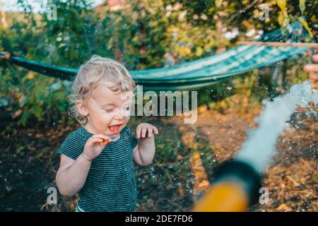 Lustige Reaktion von Baby Junge während Wasser Kampf Spiel mit Schlauch. Kleinkind spielen, abkühlen bei heißem Wetter - Sommer oder Frühjahr Saison. Stockfoto