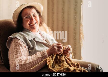 Ältere Frau lacht laut, während sie Pullover für ihre Enkelkinder strickt. Stockfoto