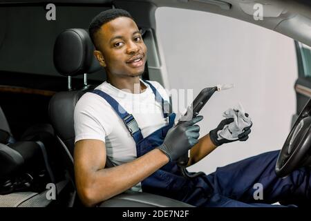 Auto Dampfreinigung, Desinfektion. Hübscher junger afroamerikanischer Kerl Auto-Service-Arbeiter, trägt weißes T-Shirt und Overalls, sitzt innen Stockfoto