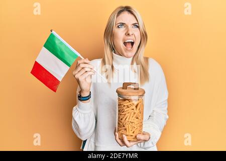 Junge kaukasische Frau hält Glas Makkaroni Pasta und italienische Flagge wütend und verrückt schreien frustriert und wütend, schreien mit Wut aufschauen. Stockfoto