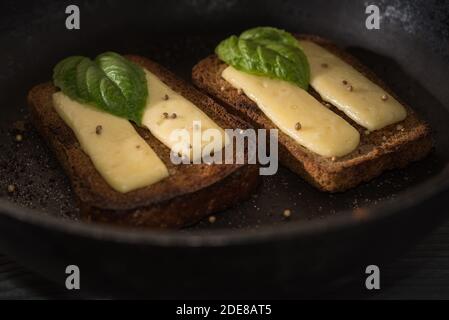 Geröstete Brotscheiben mit Käse und Koriandersamen, garniert mit frischen Basilikumblättern, in einer gusseisernen Pfanne auf schwarzem Hintergrund. Selektiver Fokus. Stockfoto