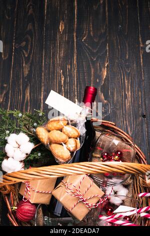 Flaschen Rotwein im Weihnachtskorb. Glühwein mit Geschenkschachteln, Keksen und Tannenbaumschmuck. Dunkler Holzhintergrund Stockfoto