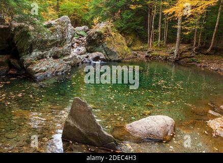 Bach, der zwischen großen Felsbrocken in einen kristallklaren smaragdgrünen Pool stürzt. Schwimmloch auf dem Mad River. Bäume ändern ihre Farbe im Herbst. Warren, VT, USA Stockfoto