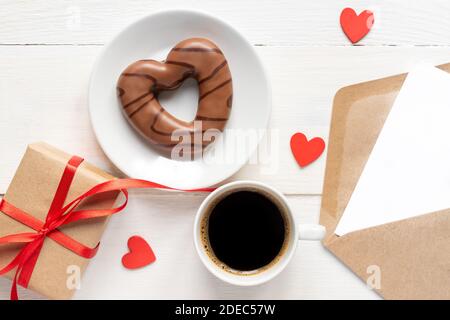 Valentinstag Mockup. Kaffee, herzförmige Schokoladenkekie, Geschenkbox und Papierkarte mit Bastelumschlag auf weißem Holztisch Draufsicht. Flacher Lay Sty Stockfoto