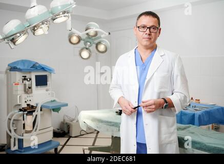 Vorderansicht des Mannes Chirurg in der Brille Blick auf die Kamera mit ernstem Ausdruck, während im Operationssaal im Krankenhaus stehen. Konzept der Medizin, medizinisches Personal und plastische Chirurgie. Stockfoto