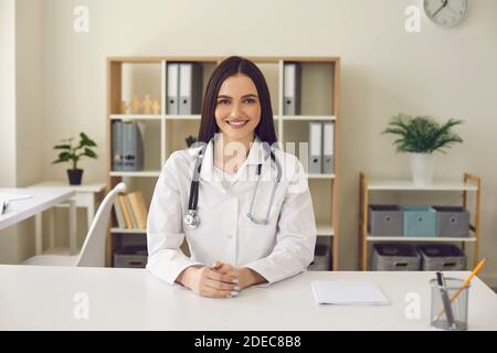 Porträt einer jungen selbstbewussten Ärztin, die am Tisch sitzt und lächelt und die Kamera anschaut. Stockfoto