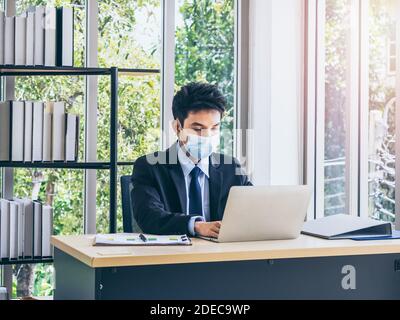 Junge gut aussehende asiatische Geschäftsmann in Anzug trägt schützende Gesichtsmaske Arbeiten auf Laptop-Computer mit Diagramm für die Überprüfung Business-Plan Während ich sitze Stockfoto