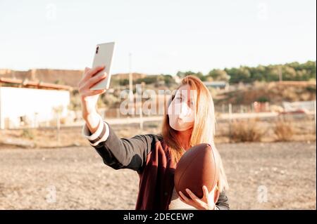 Frau trägt eine schützende Gesichtsmaske und mit ihrem Smartphone, während sie einen american Football Ball hält. Neues normales Konzept. Stockfoto