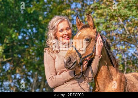 Portait einer schönen blonden Frau mit den Armen um den Hals ihres gelben Fohlens. Im Herbst Sonnenlicht, Bäume im Hintergrund. Stockfoto