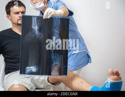 Ärztin in einem blauen medizinischen Gewand erklärt einem männlichen Patienten mit gebrochenem Bein das Ergebnis einer Röntgenaufnahme. Stockfoto