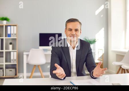 Porträt eines glücklichen Geschäftsmannes, der am Schreibtisch im Büro sitzt, die Kamera anschaut und spricht Stockfoto