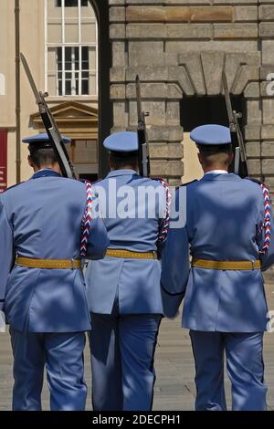 Rückansicht von drei Wachposten der Prager Burggarde in ihren blassblauen Sommeruniformen, die mit geschulterten Gewehren mit Bajonetten, die während einer Wachwechsel-Zeremonie im ersten Burghof fixiert wurden, vom Betrachter wegmarschieren. Die Wachposten an den Toren der mittelalterlichen Burg wechseln zur vollen Stunde, und es gibt auch einen täglichen 12 Uhr mittags zeremoniellen Wachwechsel, einschließlich einer Fanfare und einer Flaggenzeremonie. Das Schloss ist der offizielle Sitz und das Büro des Präsidenten der Tschechischen Republik oder Tschechiens, und die Hauptaufgabe der Wachen ist es, den Präsidenten zu schützen. Stockfoto