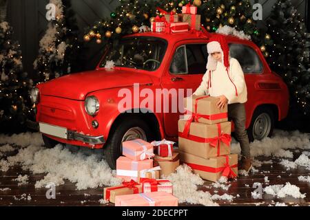 Junge auf dem Hintergrund eines roten Autos mit Geschenken. Kind baut einen Turm aus Weihnachtskisten. Stockfoto