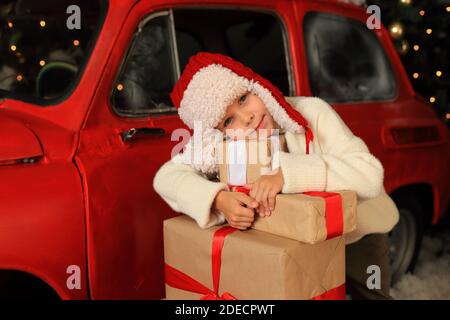 Junge auf dem Hintergrund eines roten Autos mit Geschenken. Kind baut einen Turm aus Weihnachtskisten. Stockfoto