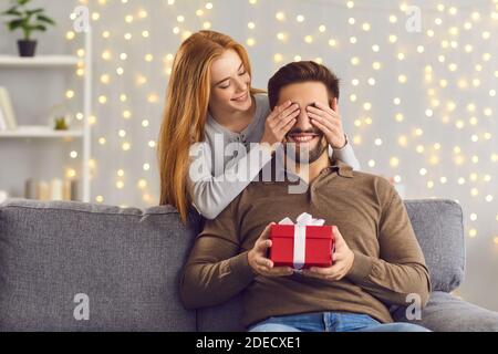 Lächelnde junge Frau bedeckt die Augen ihres Freundes und gibt ihm Ein Überraschungsgeschenk Stockfoto