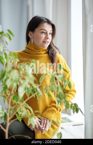 Ein Mädchen in einem gelben Pullover sitzt in frischen grünen Blättern und blickt aus dem Fenster. Frühlingsfrische und Schönheit. Stockfoto