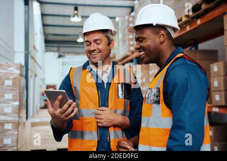 Multiethnische Fabrikingenieure lächeln beim Blick auf den digitalen Tablet-Bildschirm Im Industrielager Stockfoto