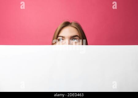 Schönes junges Mädchen, das hinter einer weißen Papierwand auf einem rosa Hintergrund herausguckt, isoliert Stockfoto