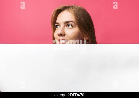 Schönes junges Mädchen, das hinter einer weißen Papierwand auf einem rosa Hintergrund herausguckt, isoliert Stockfoto