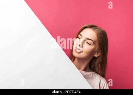 Schönes junges Mädchen, das hinter einer weißen Papierwand auf einem rosa Hintergrund herausguckt, isoliert Stockfoto