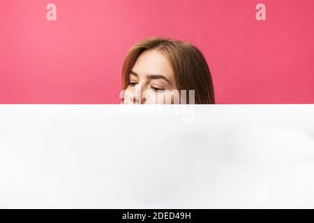 Schönes junges Mädchen, das hinter einer weißen Papierwand auf einem rosa Hintergrund herausguckt, isoliert Stockfoto