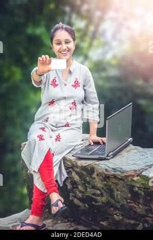 Eine schöne junge indische Dorf Frauen Online-Shopping mit ihrer Kreditkarte sitzt vor ihrem ländlichen Haus. Herbstwaldlandschaft. Stockfoto