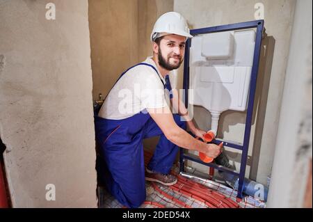 Schöner bärtiger Mann in der Arbeit Overalls Installation verdeckter Toilettenrahmen im Badezimmer. Fröhlicher männlicher Arbeiter im Schutzhelm, der auf die Kamera schaut und lächelt, während er ein an der Wand hängende Toilettensystem installiert. Stockfoto