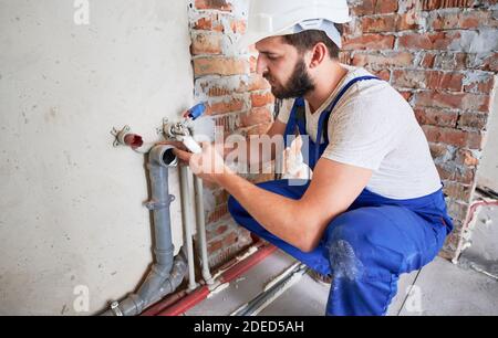 Junge Klempner, trägt blaue Uniform und weißen Helm arbeiten mit Dichtstoff fix von Kanalrohr in der Küche oder im Badezimmer in unfertigen Wohnung. Konzept der Sanitär-Arbeiten und Renovierung Stockfoto