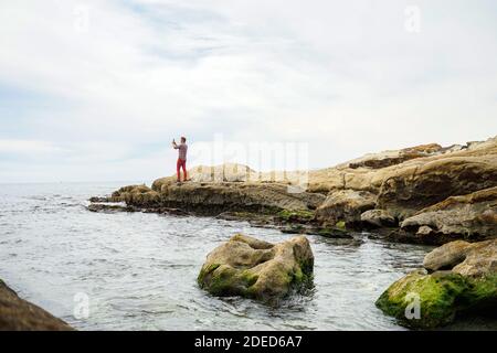 Reisender Mann fotografieren mit dem Smartphone vom Meer felsigen Ufer, nebligen Tag Stockfoto