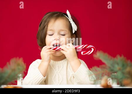 Kleines Mädchen 2-4 in gestricktem Kleid versucht Lollipop mit geschlossenen Augen auf rotem Hintergrund. Frohe weihnachten und Frohes neues Jahr. Stockfoto