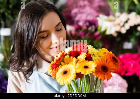 Brunette Floristin mit geschlossenen Augen riechende Gerberas in Vase in der Nähe Verschwommene Blumenvielfalt auf dem Hintergrund Stockfoto