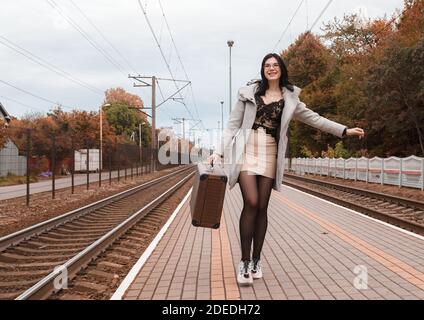 Junge lächelnde Mädchen in einem grauen Mantel mit einem Koffer Spaziergang am Herbsttag entlang des Bahnhofs Stockfoto