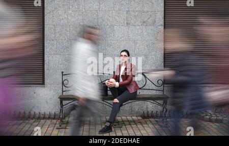 Einsames junges Mädchen in einer braunen Jacke und schwarzen Jeans Sitzen auf einer Bank in der Nähe des Bürgersteig unter einem Gleichgültigen Menge am Herbsttag Stockfoto