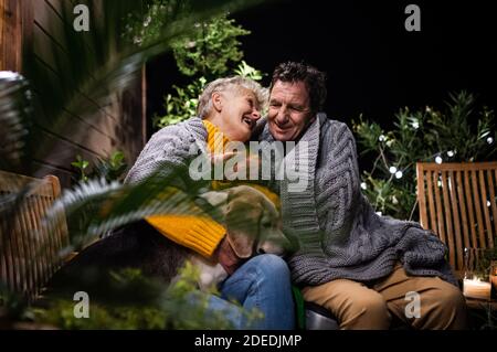 Porträt eines älteren Paares mit Hund sitzt am Abend auf der Terrasse, umarmt. Stockfoto