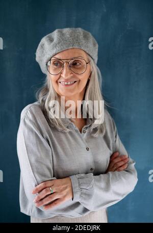 Porträt einer älteren Frau mit grauer Baskenmütze, die vor dunklem Hintergrund im Innenbereich steht. Stockfoto