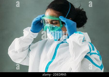 Junge weibliche Sanitäter trägt einen Schutzanzug und Maske Putting Auf der Brille und nach unten schauen Stockfoto