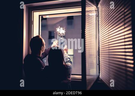 Ein Paar hält nachts Wunderkerzen aus dem Fenster. Silvesterfeier, Jubiläum, Party oder Datum zu Hause. Spontaner, ehrlicher Spaß mit Licht. Stockfoto