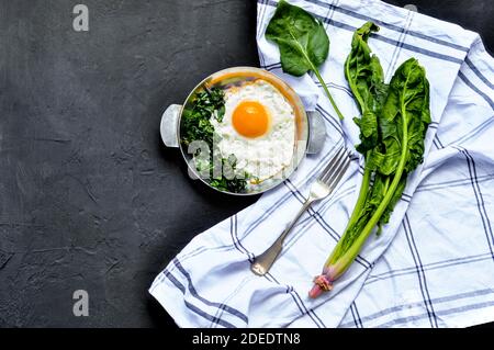 Spiegeleier mit Spinat auf eiserner Pfanne. Spiegelei Frühstück auf dunkelschwarzem Beton Hintergrund mit weißem Handtuch und Spinatblätter, Zutaten Stockfoto