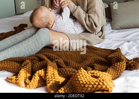 Nette kaukasische Baby Mädchen liegen auf gemütliches Bett mit ihrer Mutter in gestrickten Pullover braune Farbe gekleidet. Aufenthalt zu Hause während Coronavirus covid-19 Quarantäne Stockfoto