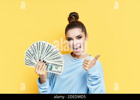 Glücklich aufgeregt junge Frau zeigt Geldscheine mit Daumen nach oben auf gelbem Hintergrund. Motivation, Erfolgskonzept Stockfoto