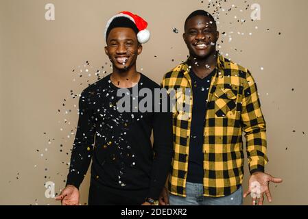 Zwei attraktive stilvolle schwarze Homosexuell Männer feiern Silvester-Party, homosexuelles Paar werfen Konfetti, gratuliert einander auf beigem Hintergrund. Weihnachten und Neujahr feiern. Stockfoto