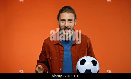 Positiver Fußballfan, der die Kamera anschaut, während er den Fußball hält Auf orange Stockfoto