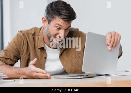 Unzufriedener Geschäftsmann gestikuliert beim Blick auf defekten Laptop am Arbeitsplatz Bei unscharfem Vordergrund Stockfoto
