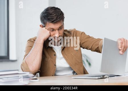 Enttäuschter Geschäftsmann, der beim Blick auf beschädigte Laptop am Arbeitsplatz denkt Bei unscharfem Vordergrund Stockfoto
