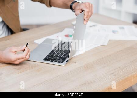 Beschnittene Ansicht des Geschäftsmannes mit Detail halten gebrochenen Laptop an Tabelle mit Papierkram auf verschwommenem Hintergrund Stockfoto